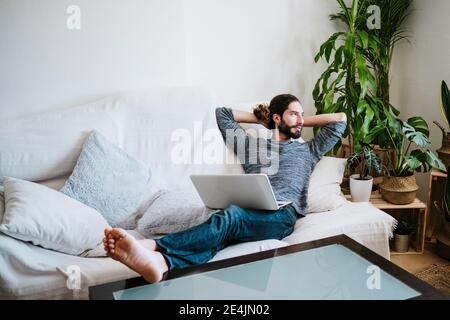 Mann mit Händen hinter dem Kopf sitzend mit Laptop auf dem Sofa Zu Hause Stockfoto
