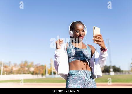Sportlerin, die Musik hört, während sie Selfie über Smartphone gegen nimmt Der Himmel ist klar Stockfoto
