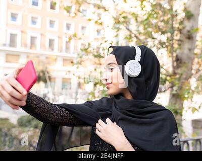 Portrait der jungen schönen Frau trägt Hijab und Kopfhörer nehmen Selfie auf dem Smartphone Stockfoto