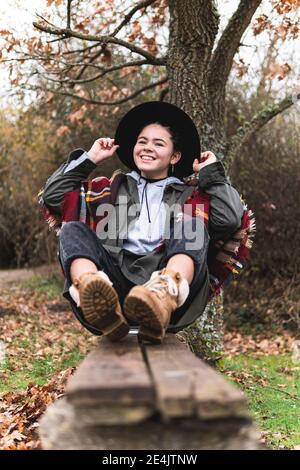 Lächelndes Teenager-Mädchen im Hut sitzt auf Planke im Herbst Querformat Stockfoto