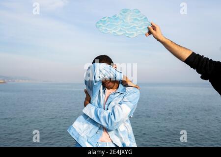 Männchen hält Wolke ausgeschnitten auf jungen Mann Kopf stehend Und das Gesicht gegen den Himmel bedecken Stockfoto