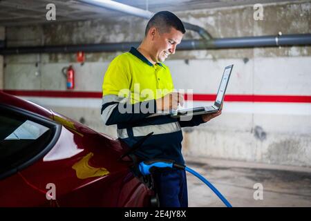 Lächelnder männlicher Programmierer mit Handy, während sich auf elektrische Auto in der Autowerkstatt Stockfoto