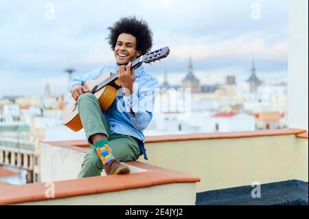 Fröhlicher junger Mann, der Gitarre spielt, während er auf der Terrasse gegen den Himmel sitzt Stockfoto