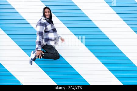 Fröhlicher junger Mann, der gegen gestreifte blau-weiße Wellpappe springt Wand Stockfoto