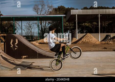 Junger Mann, der am sonnigen Tag Wheelie im Bikepark macht Stockfoto