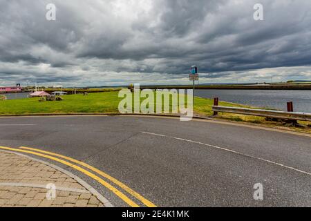 Kurve in einer leeren Straße bekannt als die lange Wanderung durch Galway Bay, grünes Gras mit dem Corrib River im Hintergrund, bewölkten Tag mit dicken grauen cl Stockfoto