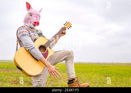 Portrait von jungen Mann mit Schwein Maske spielen Gitarre in Grasfeld Stockfoto
