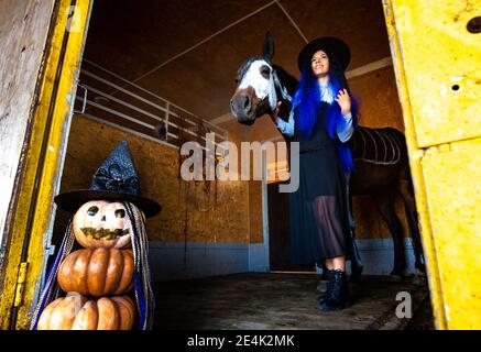 Ein Mädchen, das als Hexe gekleidet ist, kommt mit einem Pferd aus dem Corral, im Vordergrund eine böse Figur aus Kürbissen Stockfoto