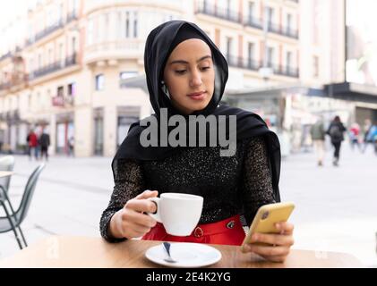 Porträt von jungen schönen Frau trägt schwarzen Hijab mit smart Telefon im Straßencafé Stockfoto