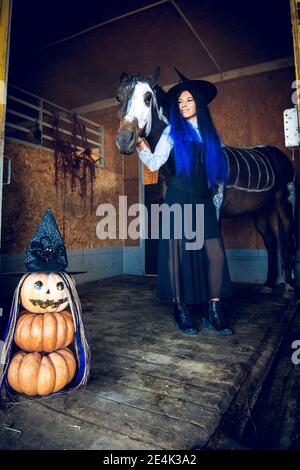 Ein Mädchen, das als Hexe gekleidet ist, umarmt glücklich ein Pferd in einem Corral, im Vordergrund eine böse Figur aus Kürbissen Stockfoto