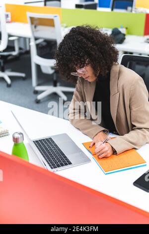 Junge Geschäftsfrau mit Laptop schreibt in Tagebuch während der Arbeit an Schreibtisch im Büro Stockfoto