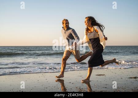 Fröhliches junges Paar, das Hände hält, während es am Strand läuft Sonnenuntergang Stockfoto