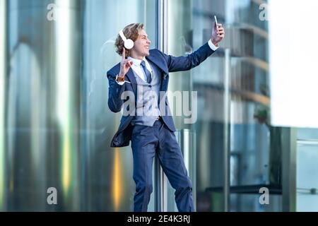 Glücklich männlich professionelle Gestik während der Aufnahme Selfie durch Smartphone Gegen Bürogebäude Stockfoto