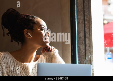 Lächelnde junge Frau, die durch das Fenster schaut, während sie mit einem Laptop sitzt Im Café Stockfoto