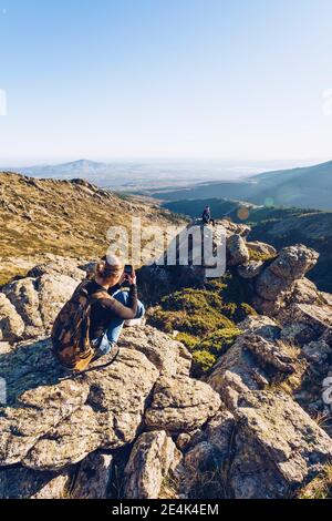 Junge Frau fotografiert männlichen Freund sitzen auf Berg gegen klar Himmel Stockfoto