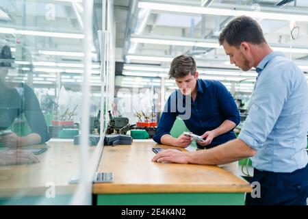 Männlicher Ingenieur, der Maschinenteil hält, mit einem Kollegen auf Schreibtisch in der Industrie Stockfoto