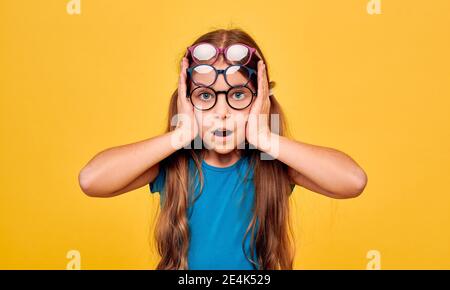 Nettes Mädchen mit überraschenden Emotionen tragen viele stilvolle Brillen auf gelbem Hintergrund. Kinder, Sehkorrektur Stockfoto