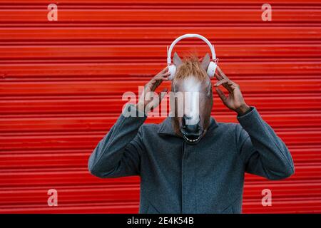 Mann mit Pferdemaske, der im Stehen Musik über Kopfhörer hört Gegen rote Wand Stockfoto