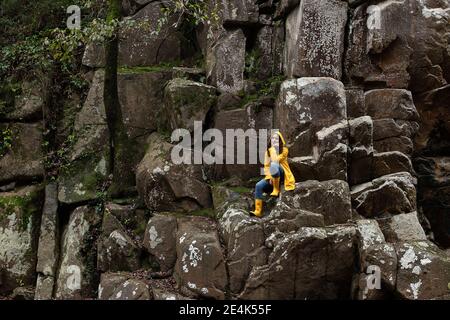 Nachdenkliche junge Frau trägt gelben Regenmantel sitzen auf Felsen in Wald Stockfoto