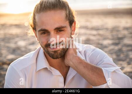 Lächelnder, hübscher junger Mann, der sich während des Sonnenuntergangs am Strand entspannt Stockfoto