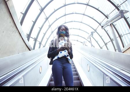 Geschäftsfrau trägt eine schützende Gesichtsmaske mit dem Telefon, während sie auf steht Rolltreppe Stockfoto