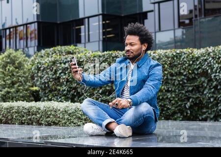 Mittelerwachsener Mann mit Afro-Haaren, der Selfie über Smart nimmt Telefonieren, während Sie draußen sitzen Stockfoto