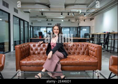 Asiatische Frau Unternehmer zuversichtlich sitzen und lächeln auf Vintage-Sofa Im Coworking Space im modernen Büro Stockfoto