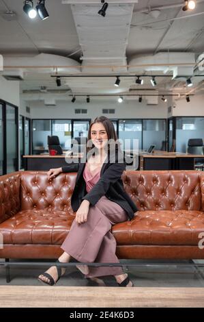 Asiatische Frau Unternehmer zuversichtlich sitzen und lächeln auf Vintage-Sofa Im Coworking Space im modernen Büro Stockfoto