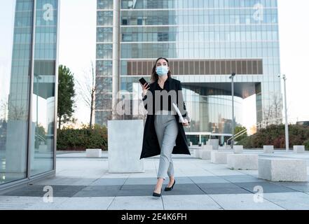 Geschäftsfrau trägt Gesichtsmaske mit Handy beim Gehen auf Fußweg Stockfoto