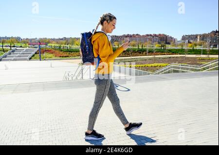 Weibliche Athletin mit Rucksack, die das Mobiltelefon benutzt, während sie auf dem Fußweg läuft Stockfoto