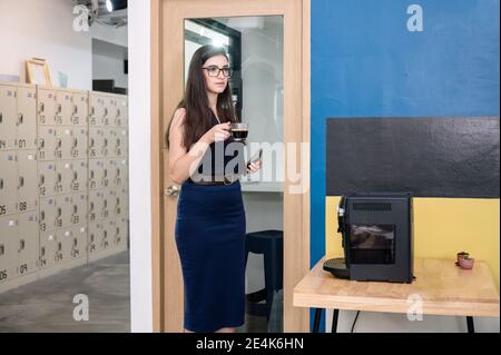Attraktive Geschäftsfrau steht mit heißen Kaffee trinken im modernen Büro Stockfoto