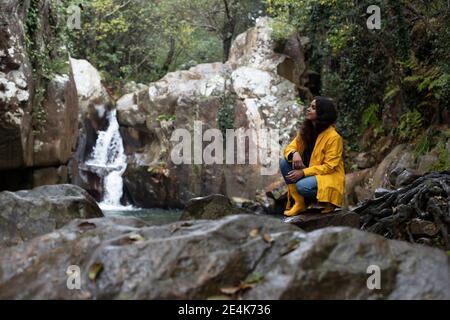 Nachdenkliche Hikerin, die auf Felsen gegen Wasserfall im Wald hockt Während der Regenzeit Stockfoto