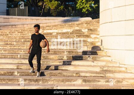 Junger Mann mit Basketball, der sich während des Sonnenuntergangs auf Stufen nach unten bewegt Stockfoto