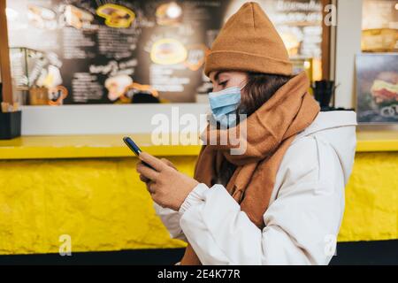 Junge Frau, die im Winter vor dem Café mit dem Smartphone wartet Während COVID-19 Stockfoto