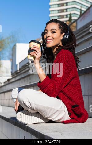 Lächelnd junge weibliche Millennial sitzend mit gekreuzten Beinen mit wiederverwendbarer Kaffeetasse An sonnigen Tag Stockfoto