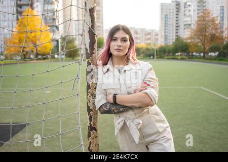 Selbstbewusste junge Frau mit gekreuzten Armen lehnt auf Goalpost in Fußballplatz Stockfoto