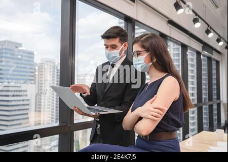 Junge kaukasische Geschäftskollegen tragen Gesichtsmaske diskutieren mit Laptop in neuen normalen Büro im Geschäftsviertel. Während Pandemie des Coronavirus, Stockfoto