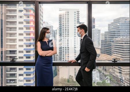 Junge kaukasische Geschäftskollege trägt Gesichtsmaske mit Gespräch durch Das Fenster im neuen normalen Büro im Geschäftsviertel Stockfoto