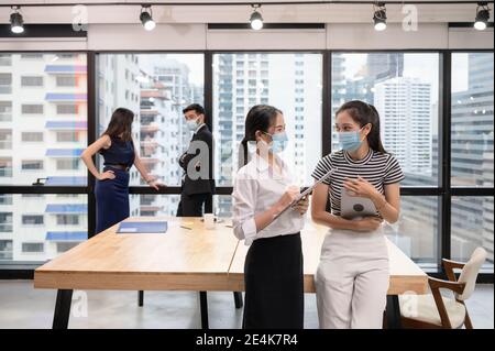 Junge asiatische Geschäftsfrau Kollegen trägt Gesichtsmaske und im neuen normalen Büro reden, kaukasische Exekutive diskutieren auf hinter im Geschäftsviertel Stockfoto