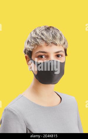 Vertikales Porträt einer zeitgemäßen kurzhaarigen Frau, die im Stehen eine Maske trägt Vor knallgelbem Hintergrund Stockfoto