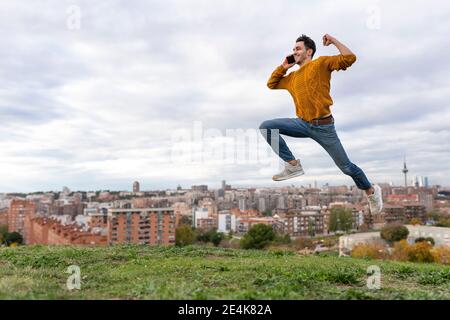 Aufgeregt junger Mann auf Handy reden, während springen über Hügel gegen Stadtbild Stockfoto