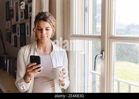 Junge schwangere Frau mit Smartphone am Fenster zu Hause Stockfoto