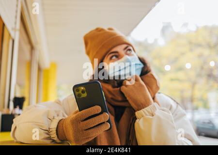 Junge Frau hält Handy und betrachtet mit der Hand am Kinn Mit dem Cafe während der COVID-19 Stockfoto