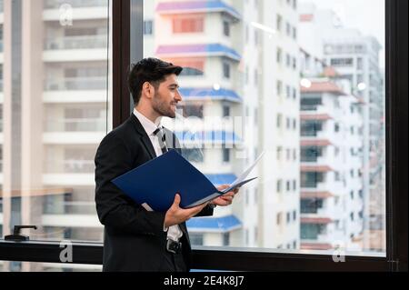 Attraktive kaukasischen Geschäftsmann hält Finanzdokument mit einem Lächeln und suchen Durch das Fenster in einem modernen Büro im Geschäftsviertel Stockfoto