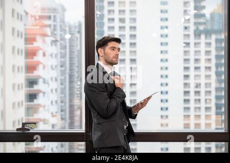 Junge kaukasische Geschäftsmann hält Krawatte und Tablet in modernen Büro Im Geschäftsviertel Stockfoto