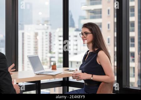 Attraktive kaukasische Geschäftsfrau sitzen mit halten Kaffeetasse und hören Kollege am Schreibtisch im modernen Büro Stockfoto
