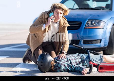 Junger Mann ruft Hilfe auf Handy mit Junge in Hintergrund liegt auf der Straße nach einem Autounfall Stockfoto