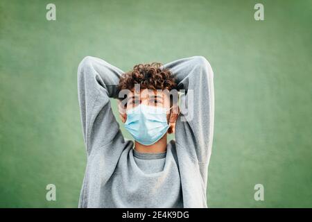 Kaukasischer Mann mit Händen hinter dem Kopf tragen chirurgische Maske gegen Grüner Hintergrund Stockfoto