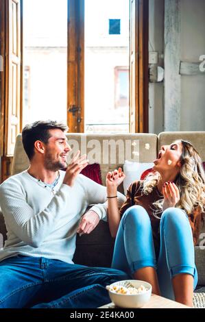 Fröhliches junges Paar, das zu Hause sitzt und mit Popcorn spielt Stockfoto