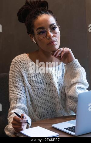 Nachdenkliche junge Frau, die im Tagebuch schreibt, während sie mit einem Laptop sitzt Im Café Stockfoto